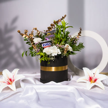 Elegant flower arrangement in black and gold box with Fox's biscuits and lilies, on white satin