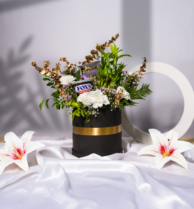 Elegant flower arrangement in black and gold box with Fox's biscuits and lilies, on white satin