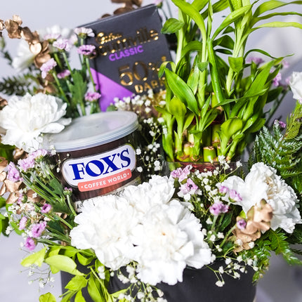 Gift basket with white flowers, green plants, Fox's Coffee World can, and dark chocolate bar