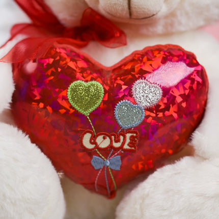 White teddy bear with red bow holding shiny red heart pillow with glitter heart balloons and 'LOVE' text