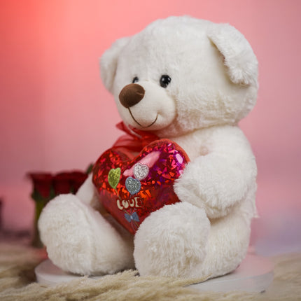 White teddy bear holding red heart pillow with 'LOVE' text, roses in background, pink backdrop