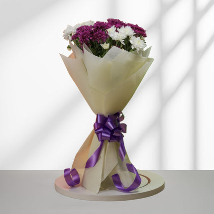 Bouquet of purple and white daisies wrapped in cream paper with a purple ribbon on a round base