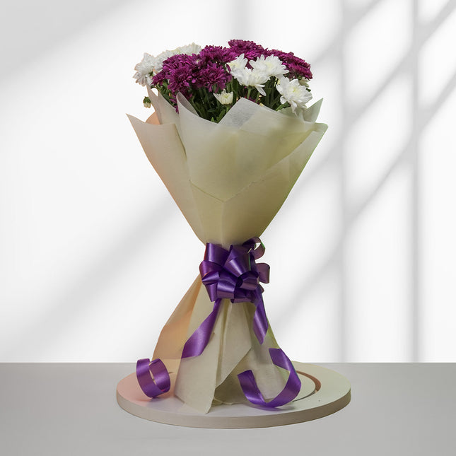 Bouquet of purple and white daisies wrapped in cream paper with a purple ribbon on a round base