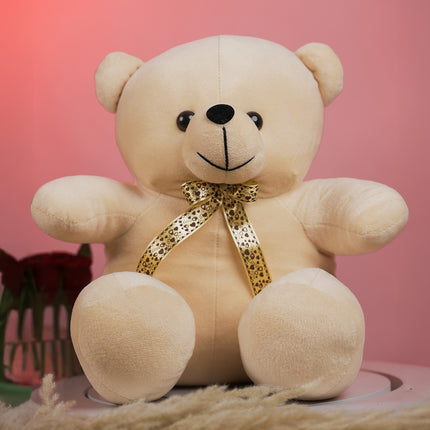 Plush beige teddy bear with gold patterned ribbon bow on pink background, roses beside