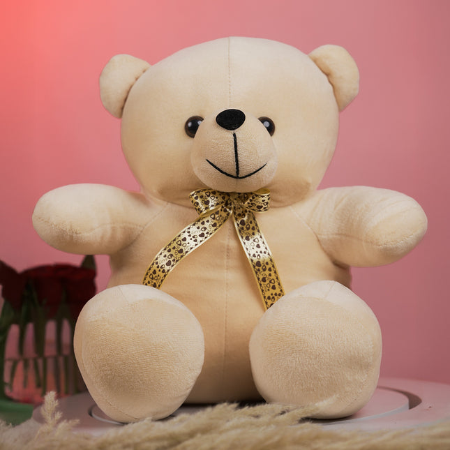 Plush beige teddy bear with gold patterned ribbon bow on pink background, roses beside