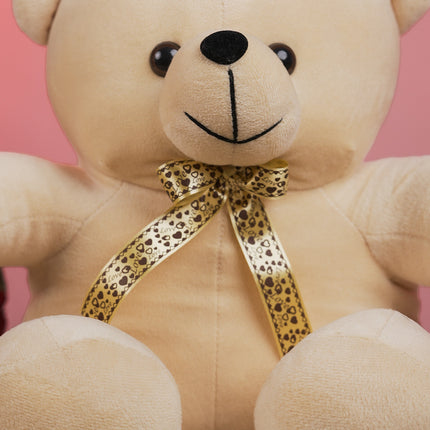 Beige teddy bear with a gold heart pattern bow sitting against a pink background