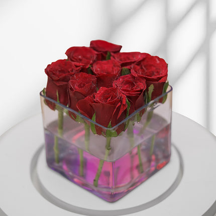 Red roses arranged in a square glass vase with pink water on a white table