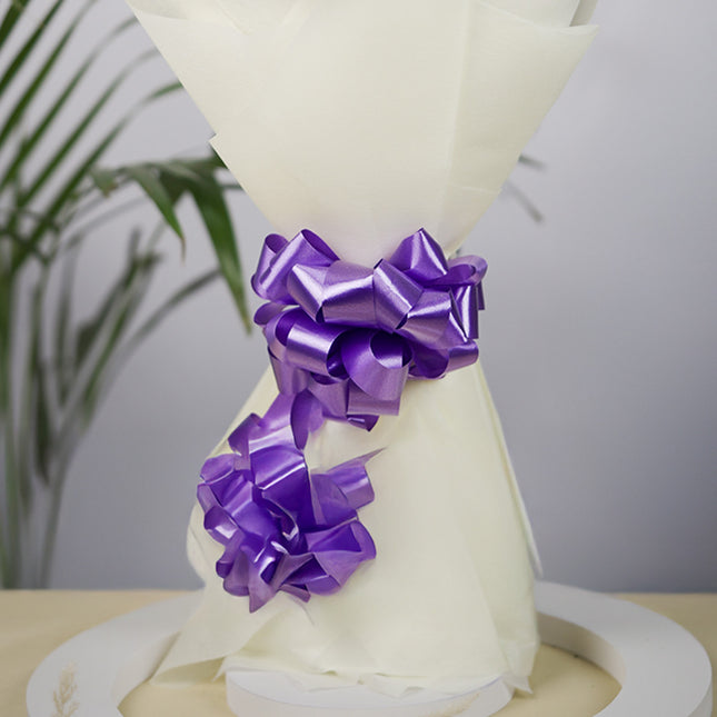 Bouquet wrapped in white paper with two shiny purple ribbon bows, indoor setting