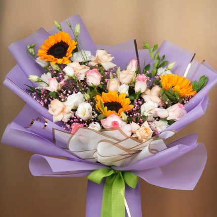 Sunflower and rose bouquet with pink accents wrapped in purple paper and green ribbon